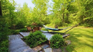 A hammock between two wicker chairs on a wooden deck, surrounded by lush greenery and a stone pathway. - Cozy, welcoming Berkshires cottage – perfect home base & place to unplug! (Stockbridge)