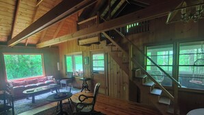A cozy cabin interior with a wooden staircase, a sofa, a dining table, and large windows offering a view of the outdoors. - Cozy, welcoming Berkshires cottage – perfect home base & place to unplug! (Stockbridge)