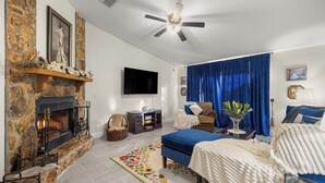 A living room with a stone fireplace, a flat-screen TV, a ceiling fan, a blue sofa, and a blue curtain. - Vintage Fall retreat Near Springs Cozy Fireplace
(Williston)