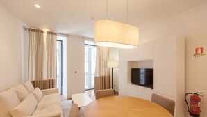 A modern hotel room with a round wooden dining table, beige sofa, and a flat-screen TV mounted on the wall. - FLH Glória 70 Flat III (Lisboa ciudad)