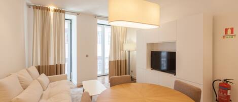 Una habitación de hotel moderna con una mesa redonda de madera, sofá color beige y un televisor de pantalla plana montado en la pared.