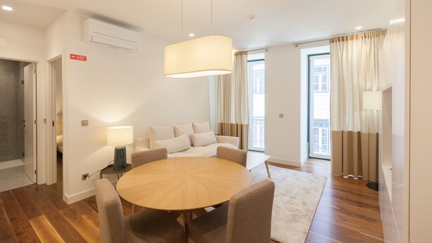 A modern living room with a round wooden table, comfortable seating, a large hanging light fixture, and a view of the outdoors through large windows. - FLH Glória 70 Flat III (Lisboa ciudad)