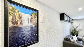 Uma sala de estar com uma grande moldura de uma cachoeira, paredes de tijolos brancos, um sofá cinza e uma estante preta com iluminação.