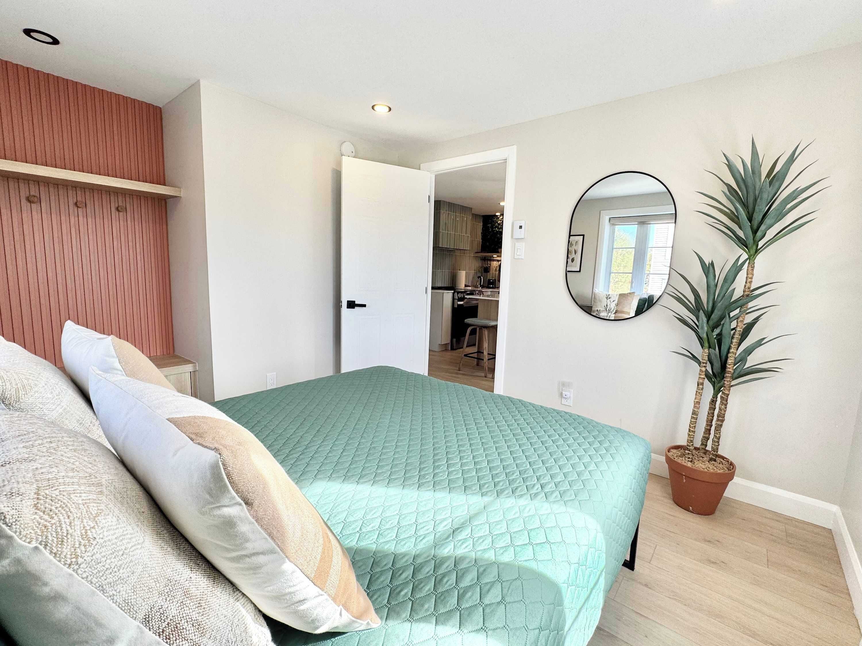 A bedroom with a green quilted bedspread, a wooden headboard, a bedside table, a potted plant, and a mirror on the wall.