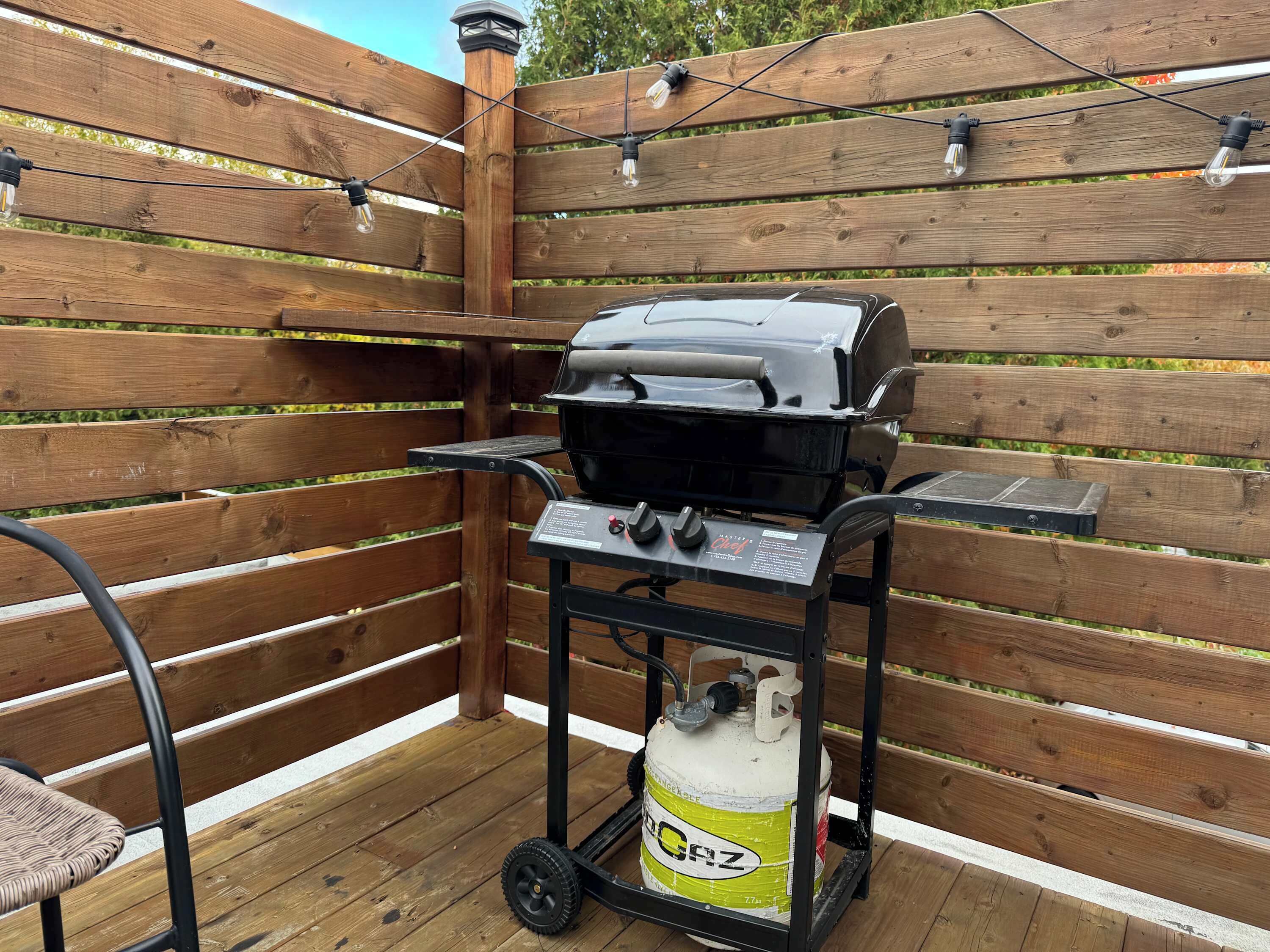 A barbecue grill with a gas tank, a metal cart, and a chair.