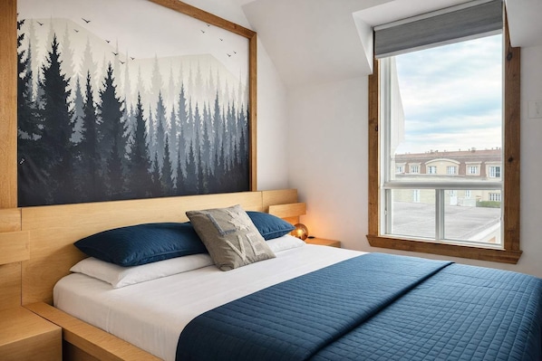 A bedroom with a bed, a painting of a forest, a window with a view, and a nightstand with a lamp.