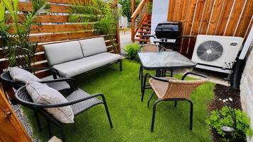 Un patio con sofá, dos sillas de mimbre, una mesa, una parrilla y un equipo de aire acondicionado.