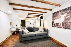 Ein modernes Wohnzimmer mit grauem Sofa, Holzbalken an der Decke, einem Bild von einem Reh und einer Küche im Hintergrund.