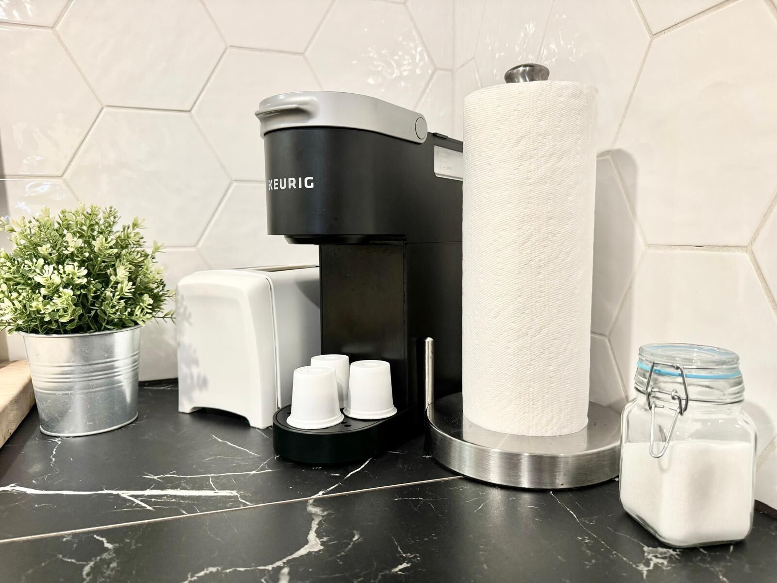 A coffee maker, a tissue roll, a small potted plant, and a jar of sugar on a kitchen counter.
