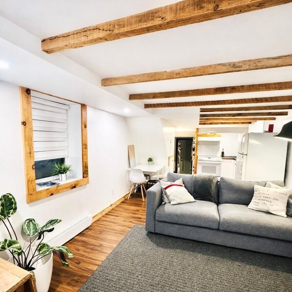 Una sala de estar moderna con un sofá gris, vigas de madera en el techo, una ventana con persianas y una pequeña mesa con plantas.