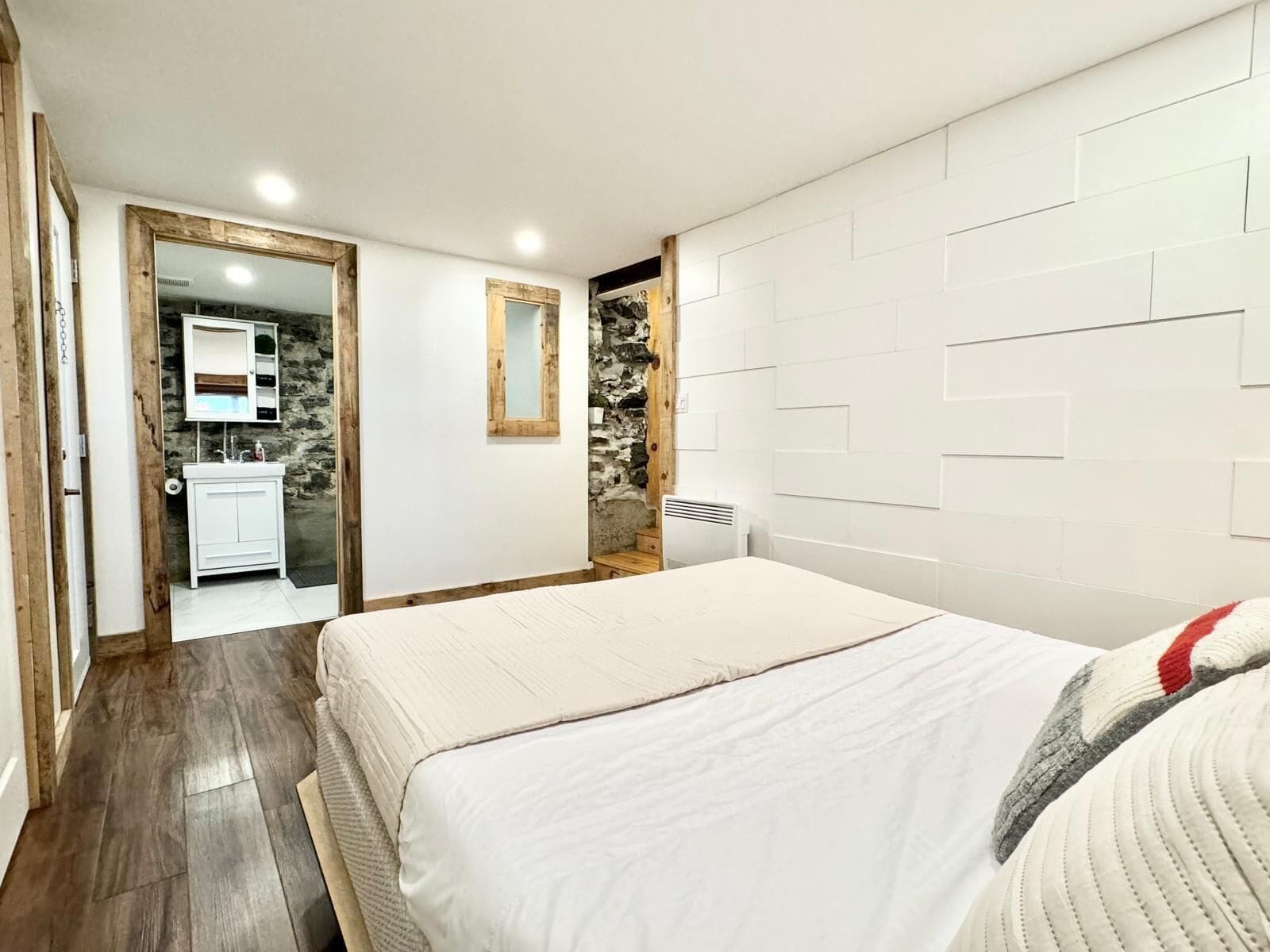A bedroom with a bed, a wooden floor, a white wall with a stone accent wall, and a bathroom with a mirror and sink.