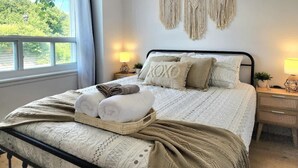 Una cama bien hecha con colcha blanca y beige estampada, dos almohadas y toallas enrolladas sobre un cabecero de madera. Hay dos mesitas de noche con lámparas, un colgador de macramé sobre la cama y una ventana con cortinas a la izquierda.