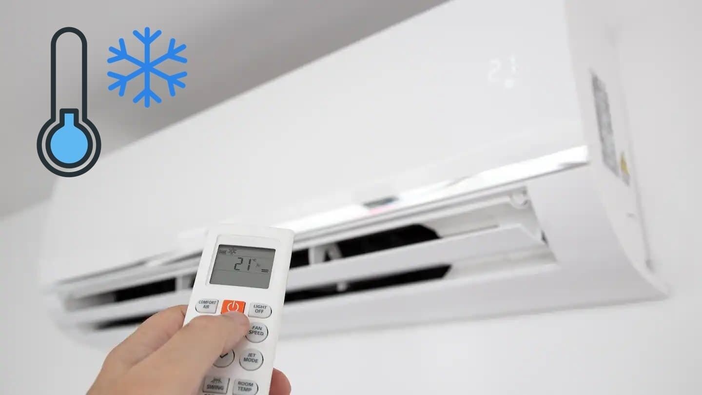 A hand holding a digital thermostat in front of a white air conditioning unit.