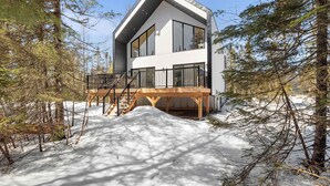Una casa moderna de dos pisos con terraza de madera y grandes ventanales, rodeada de nieve y árboles.