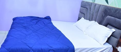 A bed with a blue quilt and white linens, a grey upholstered headboard, and a floral wall mural.