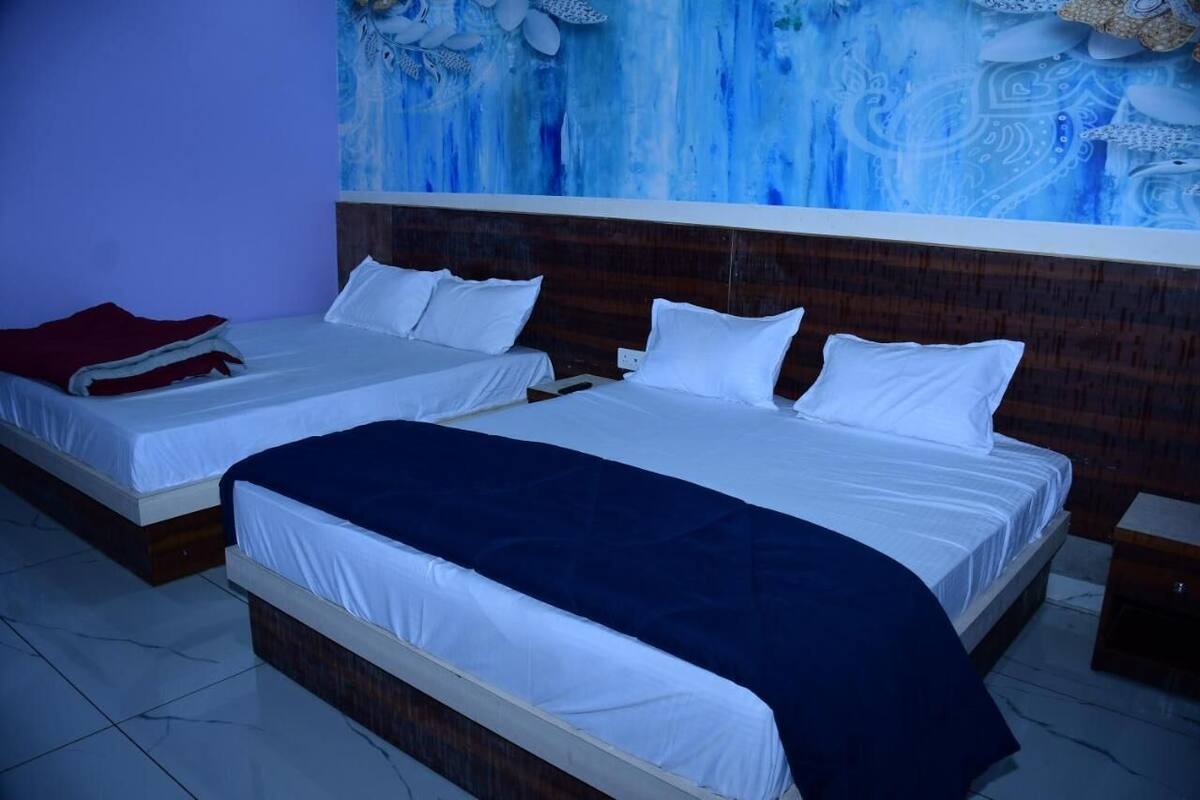 A hotel room with two beds, each with white linens and a dark blue blanket. The room has a wooden headboard and a large abstract painting on the wall.