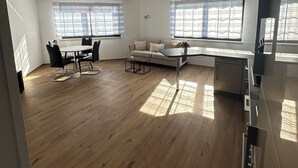 A spacious living area with a wooden floor, a sofa, a dining table with chairs, and large windows with blinds. - In & Out Boardinghouse Deluxe room (Stolberg)