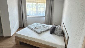 A small, modern bedroom with a single bed, a wooden nightstand, and a window with curtains. - In & Out Boardinghouse Deluxe room (Stolberg)