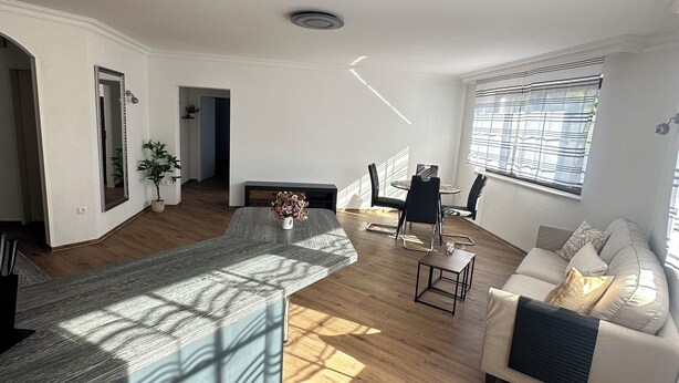 A spacious living area with a wooden floor, a sofa set, a dining table, and a large window with blinds. - In & Out Boardinghouse Deluxe room (Stolberg)