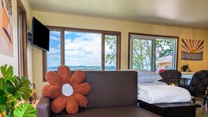 Quarto com sofá marrom, almofada grande em formato de flor, cama com roupa de cama branca, escrivaninha, duas cadeiras, TV fixada na parede e janelas amplas com vista para árvores e o céu.