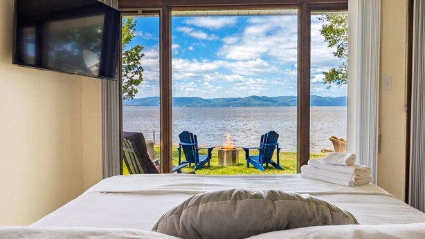Quarto com uma cama grande, TV na parede e vista para um lago com cadeiras e lareira externa.