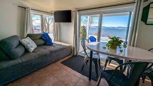 A living room with a sofa, a table with chairs, a TV mounted on the wall, and large windows with a view of the water.