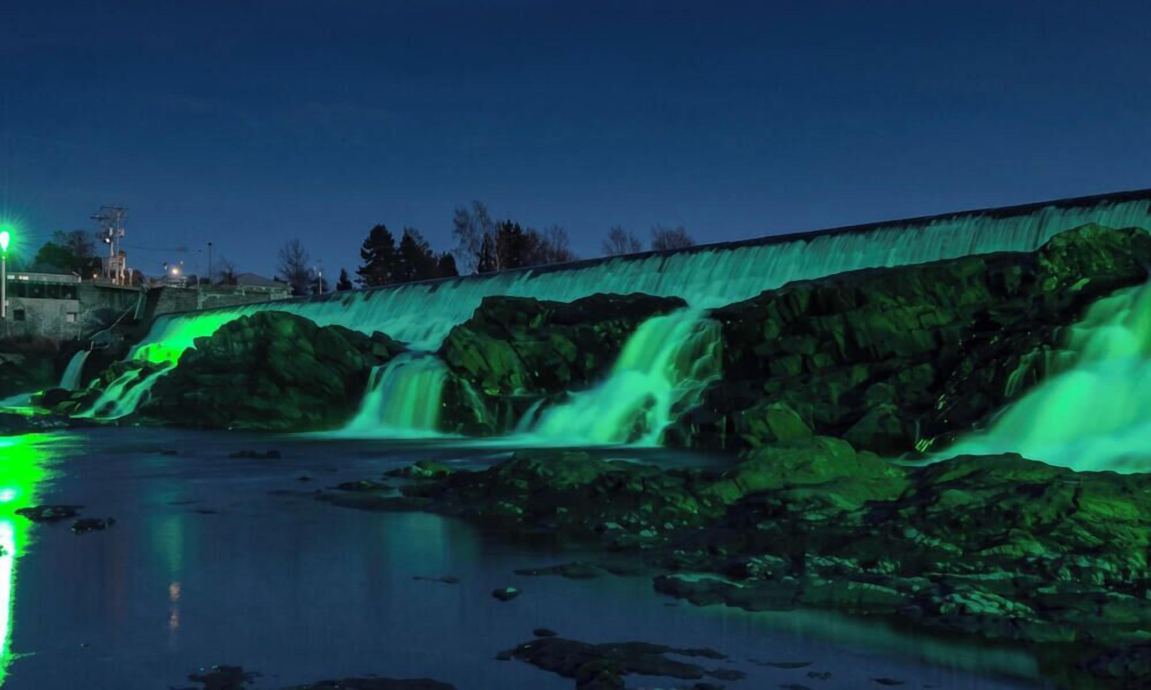 Ein Wasserfall mit grüner Beleuchtung, eine Steinmauer und ein Gebäude im Hintergrund.