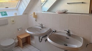 Ein modernes Badezimmer mit zwei Waschbecken, einer Toilette und einem kleinen hölzernen Hocker. Ein Oberlicht sorgt für natürliches Licht, und ein Handtuchhalter ist an der Wand zu sehen.
