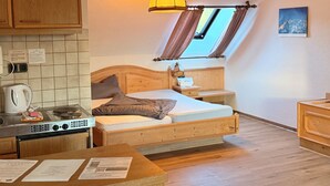Ein gemütlicher Raum mit Bett, Nachttisch, Fenster mit Vorhängen, Deckenlampe und einer Küchenzeile mit Herd und Wasserkocher.