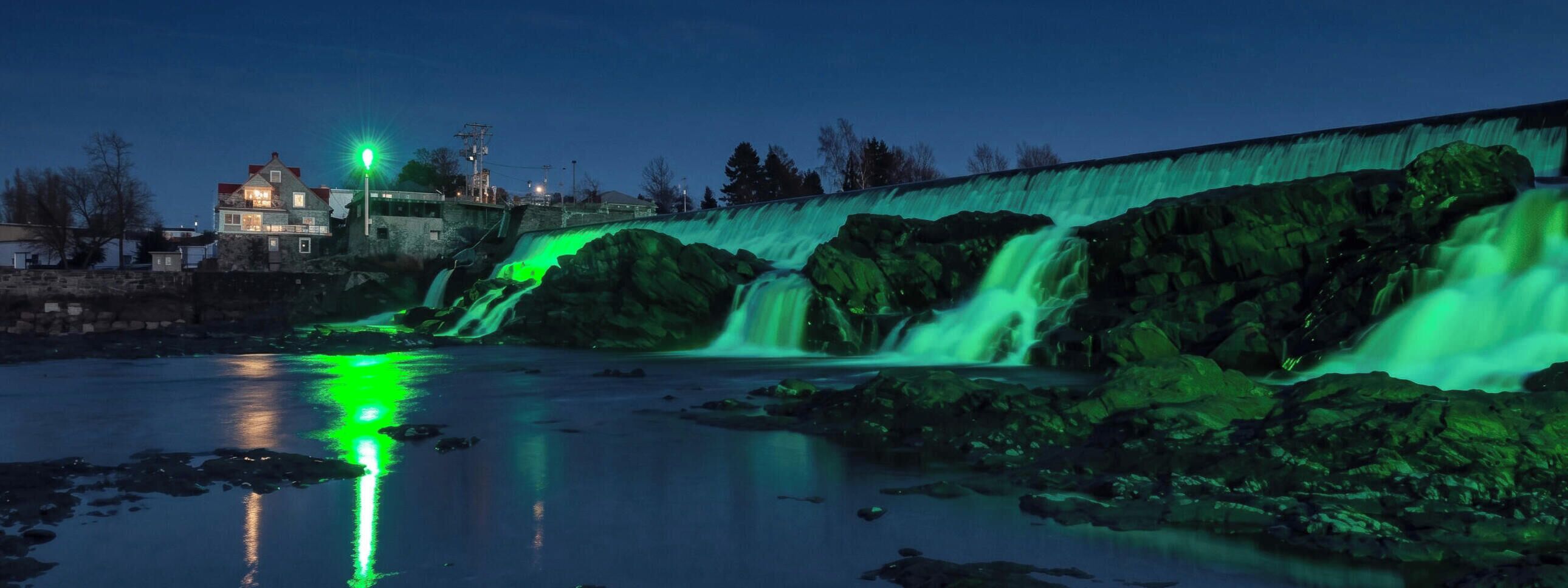 Une cascade illuminée de lumière verte, un mur de pierre et un bâtiment en arrière-plan.