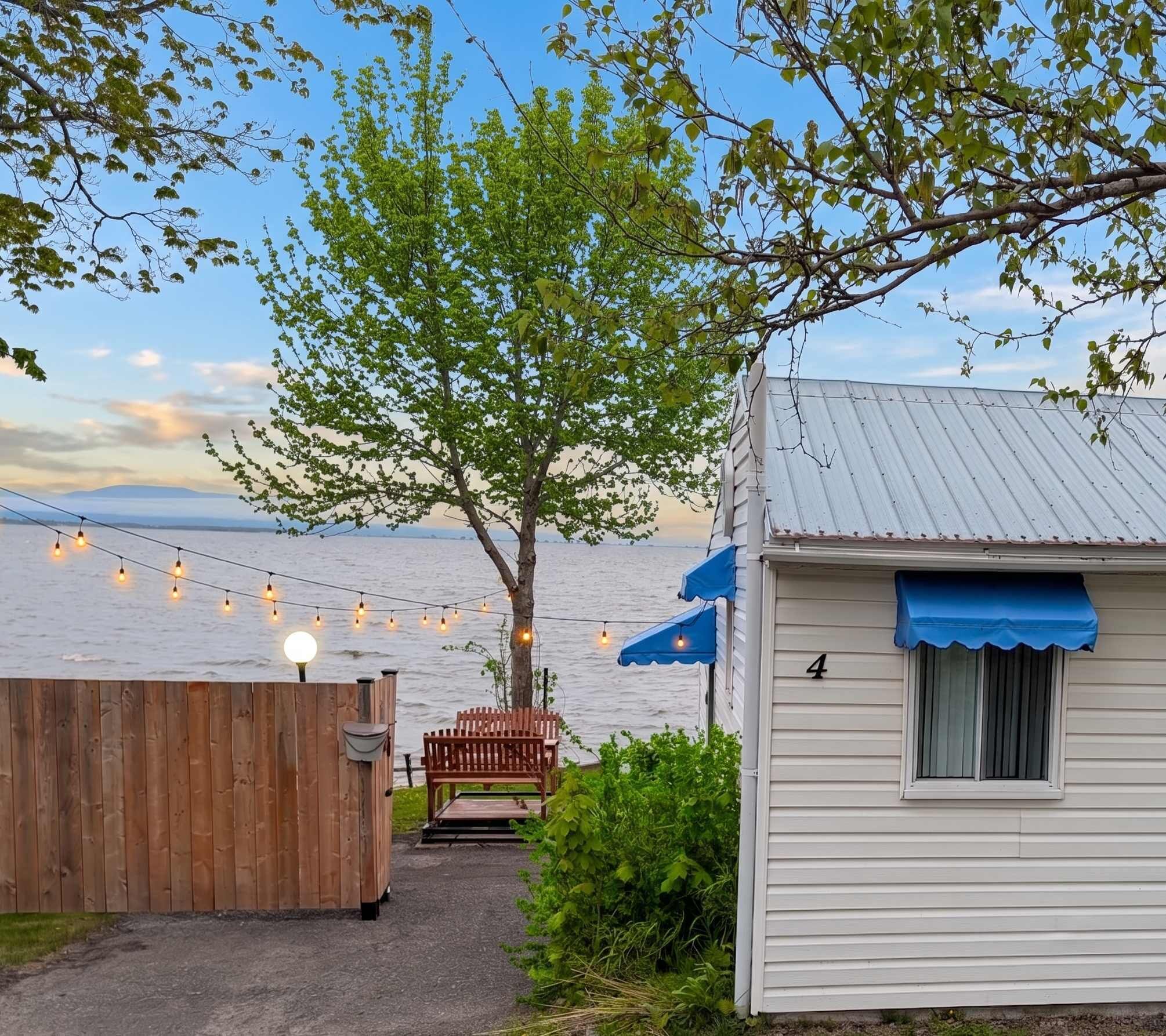 A wooden deck with a bench, a small white building with a blue awning, and string lights over a body of water.