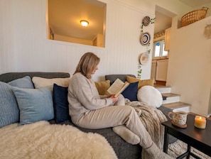Eine Person sitzt auf einem Sofa mit einem Buch, einer Tasse und einer Kerze auf einem kleinen Tisch.