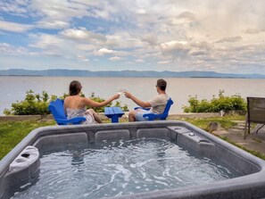 A hot tub with two people toasting, overlooking a lake with mountains in the distance.