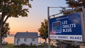 A sign for 'LES CHALETS BLEUS' with a website address, a house in the background, and a body of water. - Chalet Studio #6 Waterfront with Spa, Fireplace & Wifi (Cap-Saint-Ignace)