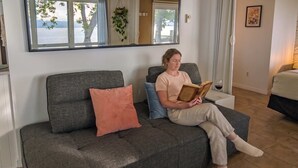 A person sitting on a grey sofa with a pink pillow, reading a book in a room with a large mirror, a window with a view of trees, and a framed picture on the wall. - Chalet #7 Waterfront with Spa, Fireplace, Wifi & AC (Cap-Saint-Ignace)