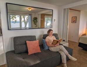 Une personne est assise sur un canapĂ© gris, avec un coussin rose, et lit un livre dans une piĂšce oĂč se trouvent un grand miroir, une fenĂȘtre donnant sur des arbres et un tableau encadrĂ© accrochĂ© au mur.