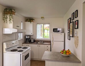 A kitchen with white cabinets, a stove, a microwave, and a sink. There is a window with a view of greenery outside. - Chalet #7 Waterfront with Spa, Fireplace, Wifi & AC (Cap-Saint-Ignace)