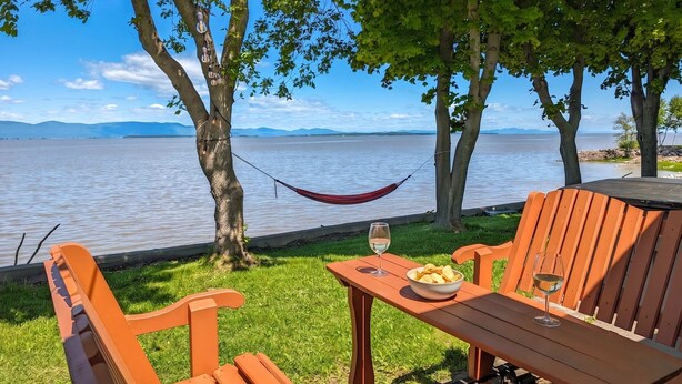 A hammock, wooden chairs, a table, and a glass of wine by a lake with trees and a clear sky. - Chalet #7 Waterfront with Spa, Fireplace, Wifi & AC (Cap-Saint-Ignace)