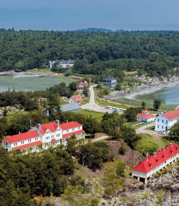 Une zone cĂŽtiĂšre comprenant plusieurs bĂątiments, dont une grande maison avec un toit rouge et un bĂątiment plus petit avec un toit gris, le tout entourĂ© de verdure et bordĂ© par une Ă©tendue dâeau.