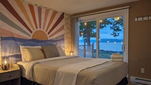 A bedroom with a large bed, a wall mural, a nightstand, a lamp, and a view of the outdoors. - Chalet #7 Waterfront with Spa, Fireplace, Wifi & AC (Cap-Saint-Ignace)