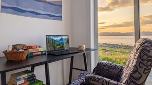A room with a laptop on a desk, a bookshelf with books, a window with a view of the sunset, and a patterned chair. - Chalet #8 Waterfront | Spa | Fireplace | Wifi | AC (Cap-Saint-Ignace)