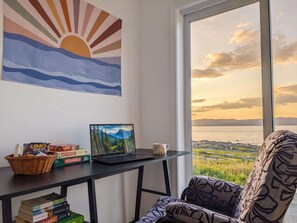 Una habitación con una computadora portátil sobre un escritorio, una estantería con libros, una ventana con vistas al atardecer y una silla estampada.