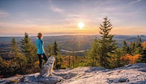 A person and a dog on a rocky outcrop overlooking a forested valley with a sunset in the background. - Chalet #9 Waterfront with Spa, Fireplace, Wifi & AC (Cap-Saint-Ignace)