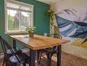 Un comedor con mesa de madera, sillas negras, una planta en maceta y un gran mural abstracto en la pared.