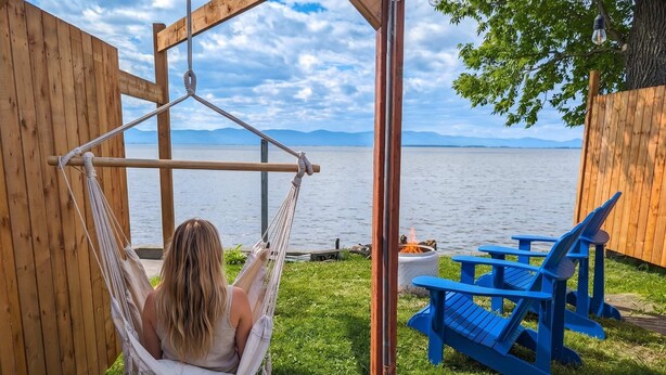 Un'amaca sotto una struttura in legno con vista su un lago, sedie blu e un braciere.