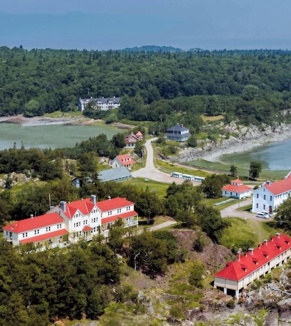 A coastal area with multiple buildings, including a large house with a red roof and a smaller building with a grey roof, surrounded by greenery and a body of water.