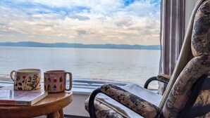 A view from a window with a chair and two mugs on a table, overlooking a body of water and distant land.