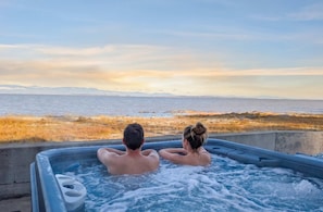 Duas pessoas em uma banheira de hidromassagem, com vista para o mar e montanhas ao longe.