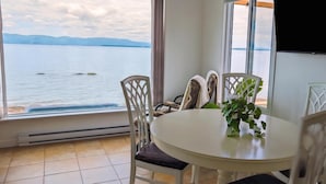 A room with a large window overlooking a body of water, a round table with chairs, and a flat-screen TV mounted on the wall.