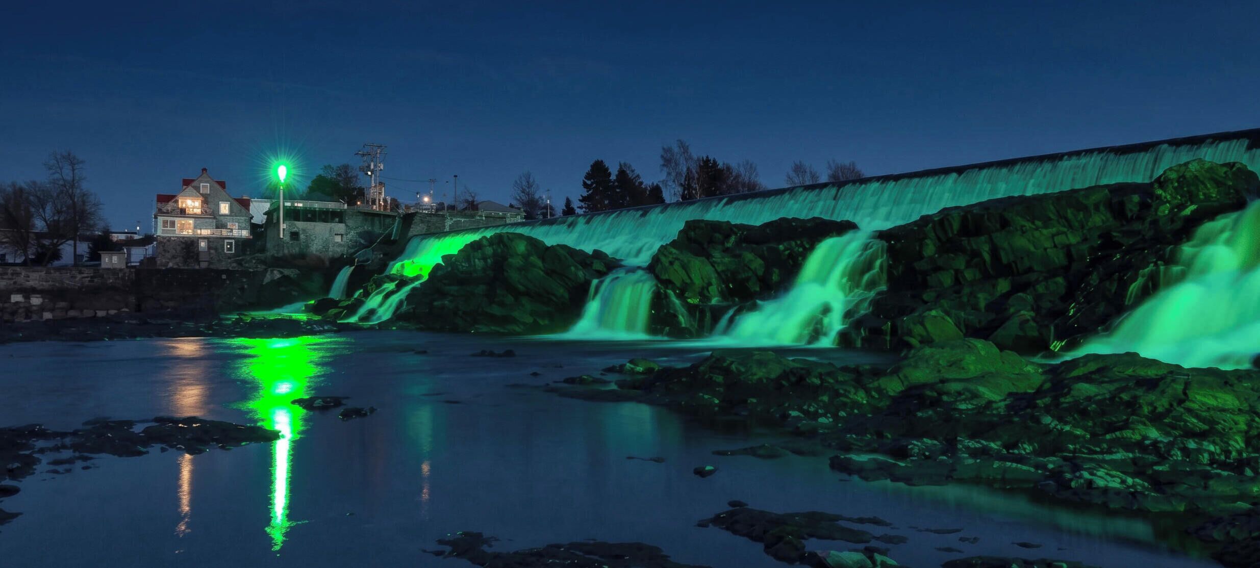 Una cascada iluminada con luz verde, un muro de piedra y un edificio al fondo.